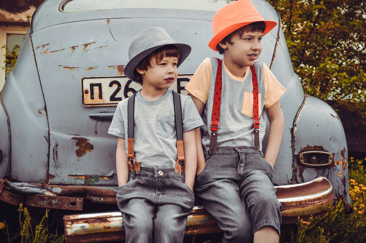 Two boys in retro-style outfits sitting on a rusty car bumper outdoors.
