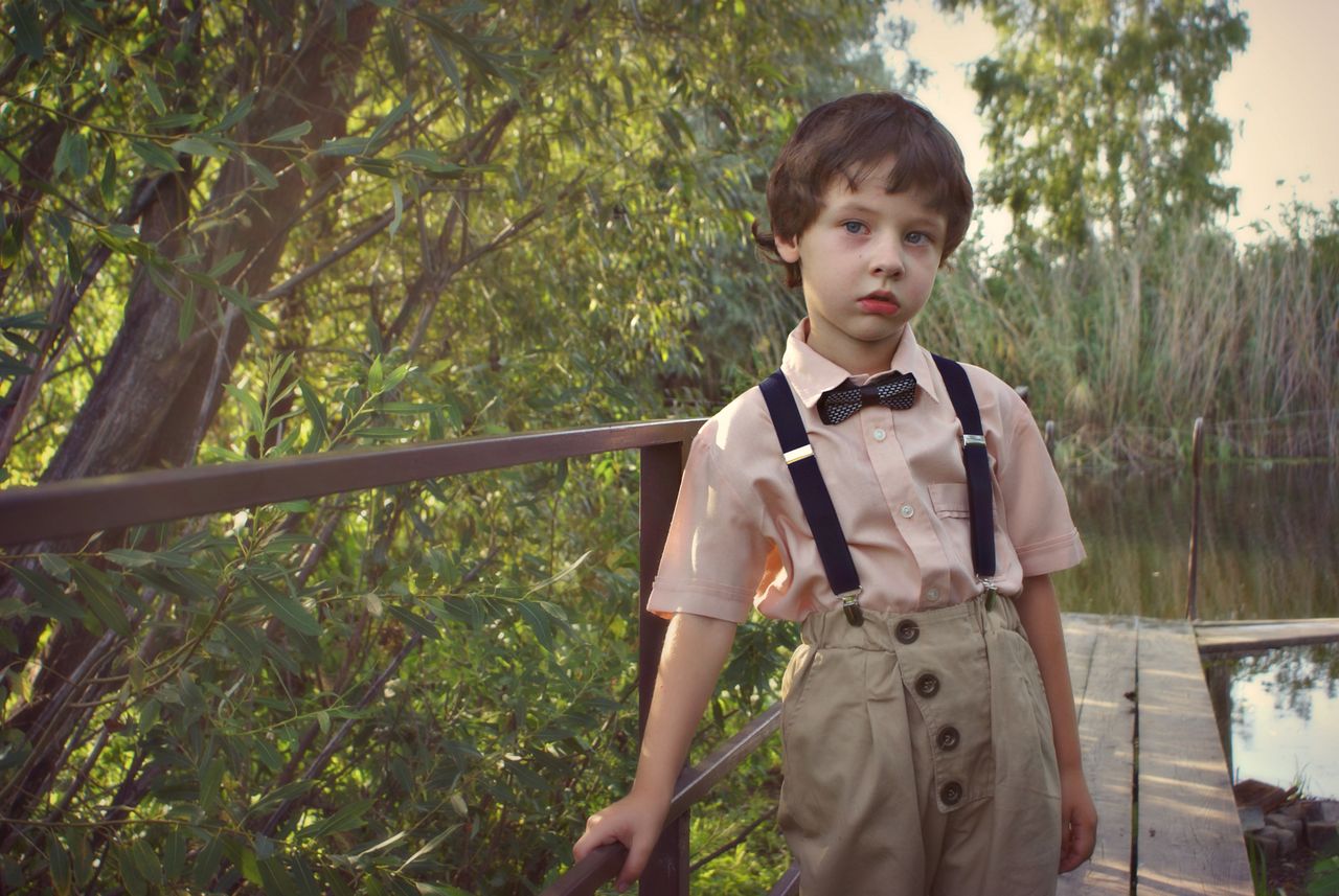 Charming young boy in vintage attire poses by a rustic bridge outdoors, showcasing classic style.