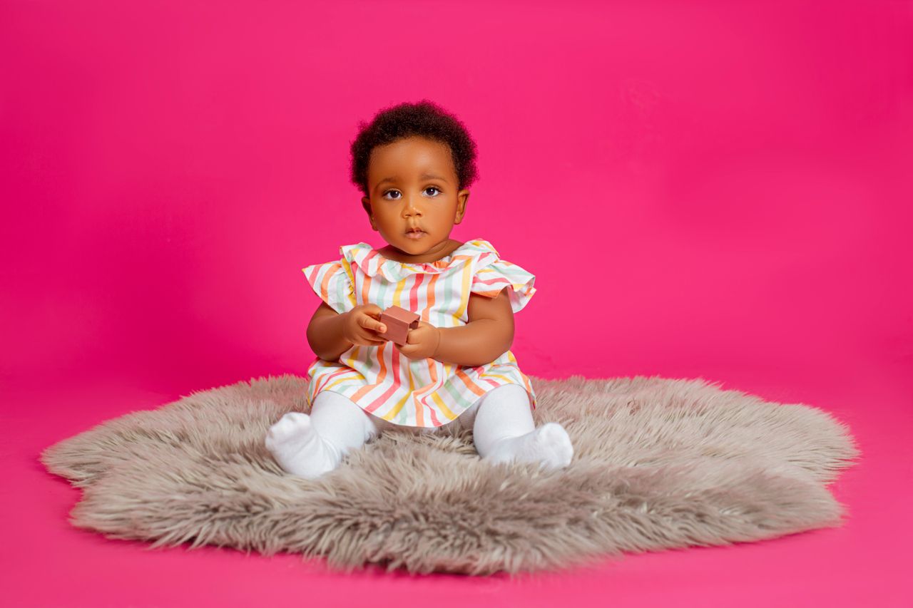 Charming baby sitting on grey rug against a bright pink backdrop, wearing colorful outfit.