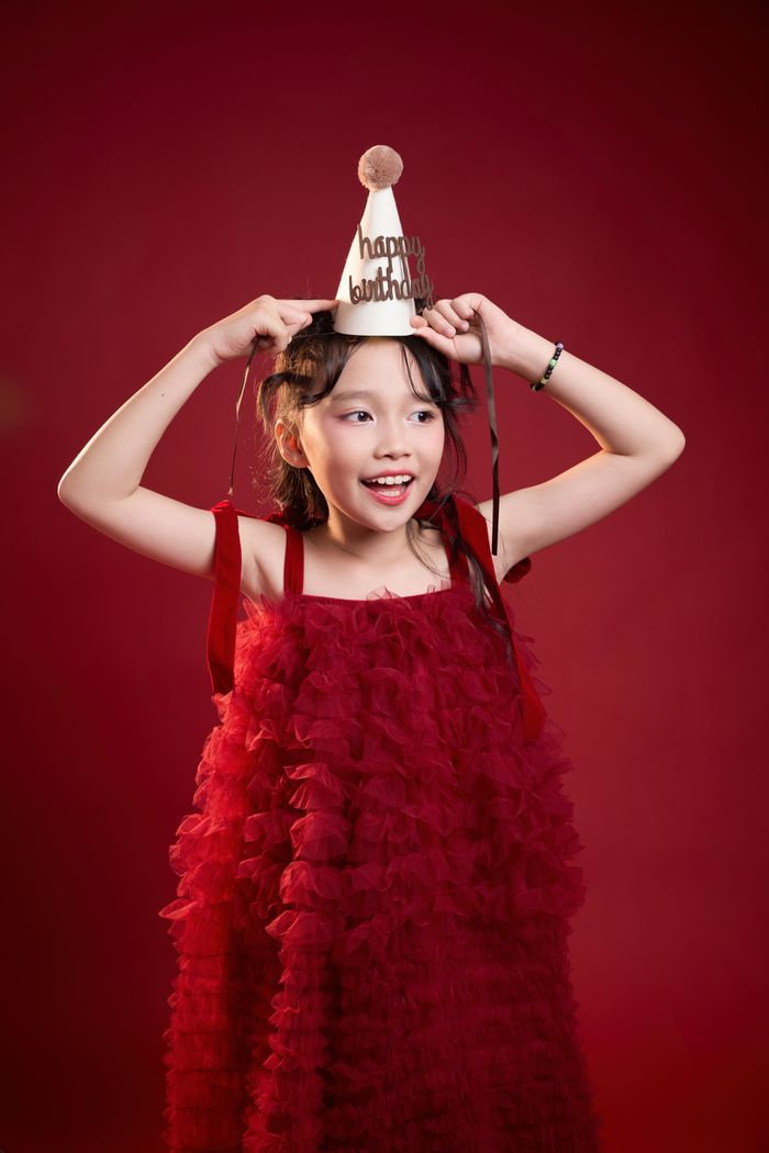 A cheerful Asian girl in a red dress celebrates her birthday with a party hat.