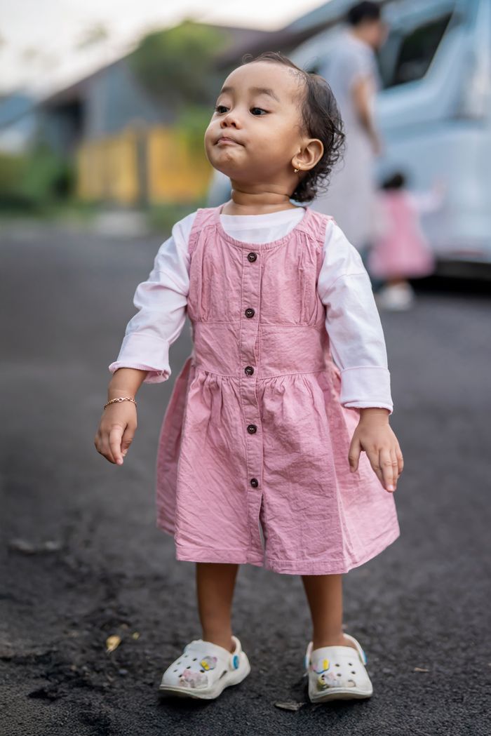 Cute toddler wearing a pink dress standing confidently outdoors with blurred background.
