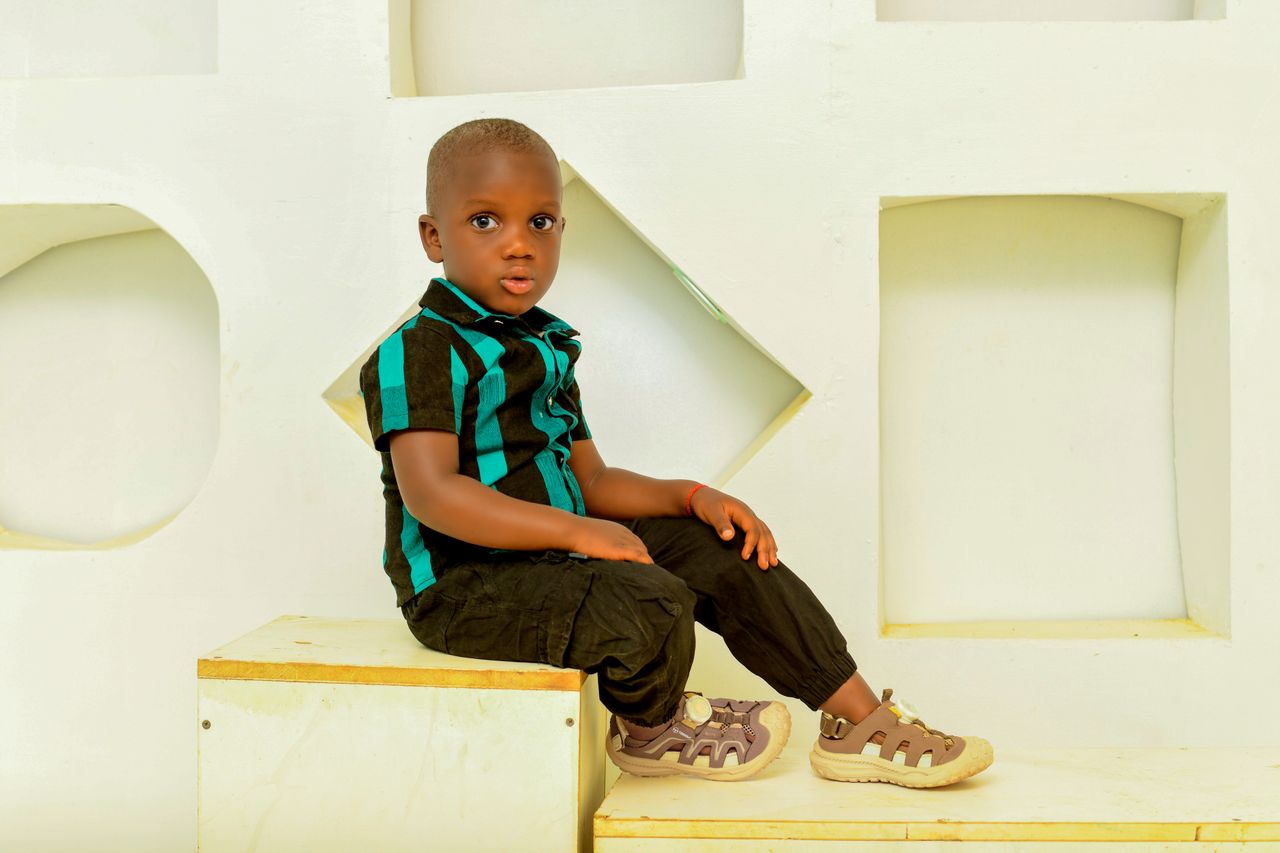 Young child sitting on a bench indoors with a stylish outfit and shoes, looking curious.
