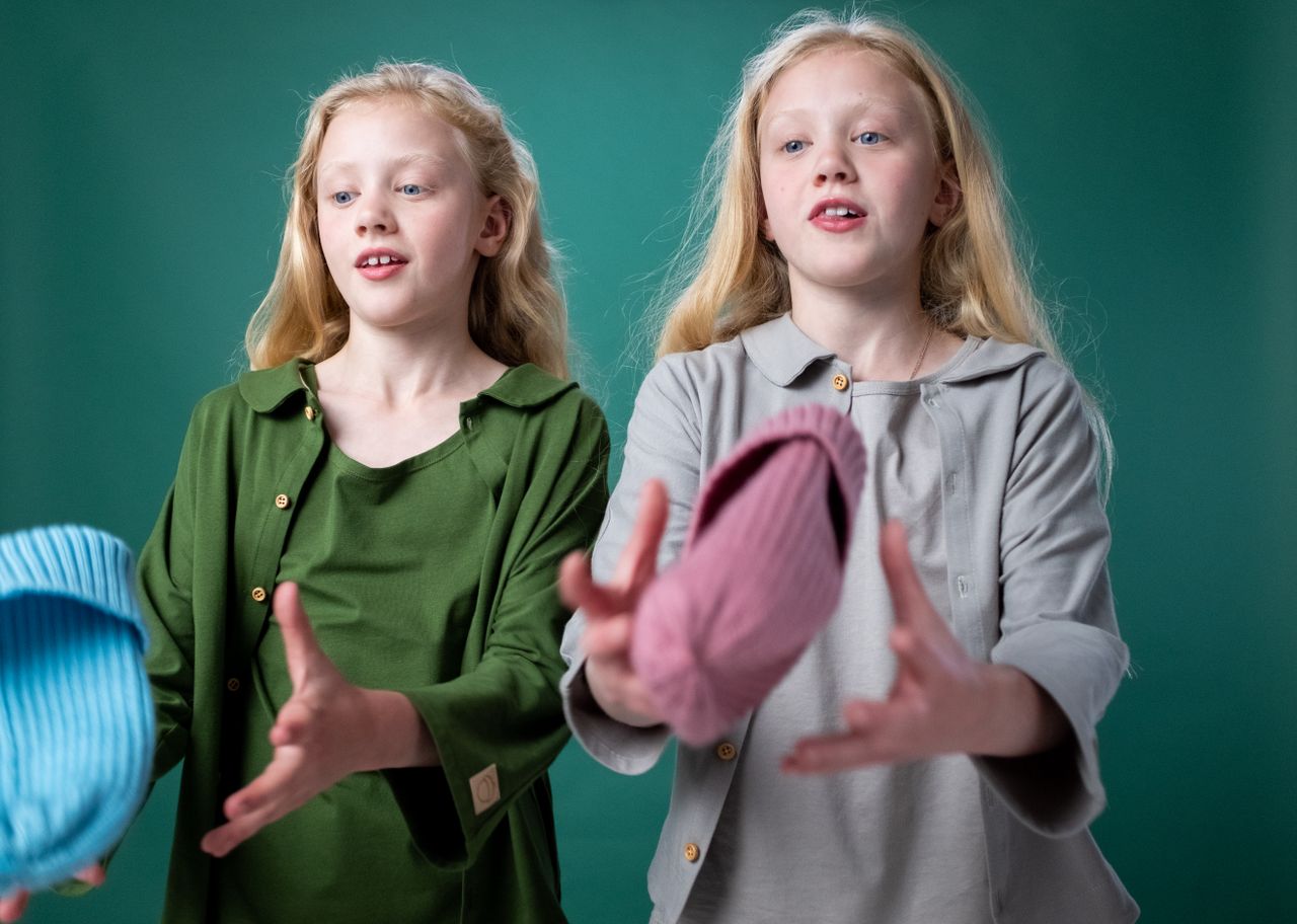 Twin girls wearing casual outfits, playfully tossing hats indoors against a green background.