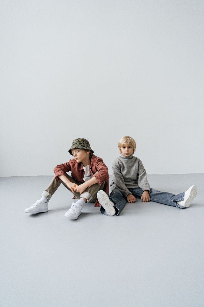 about-me-01 Two young boys in casual attire, sitting in a minimalist studio setting.