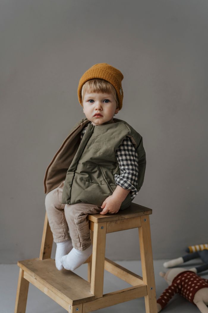 Cute child wearing knit cap and vest, sitting on a wooden chair in a studio setting.