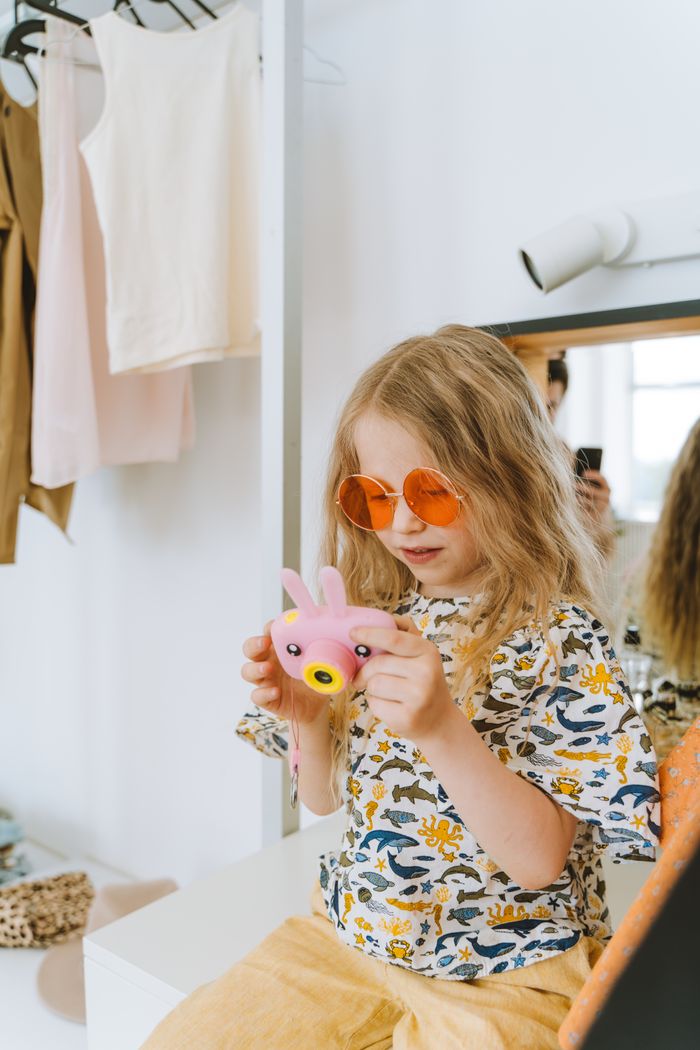 Blonde girl in sunglasses playing with a pink toy camera indoors, showcasing playful fashion.